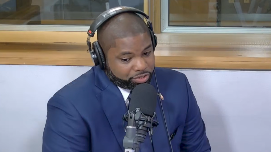 byron donalds speaks in a radio studio with a microphone in front. he is a black man wearing a blue suit
