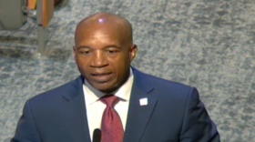 A man in a navy blue suit with a red tie speaks in front of a microphone. 