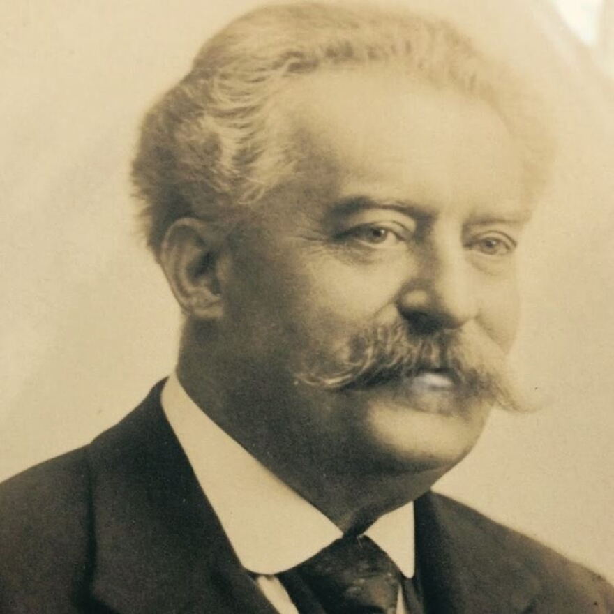 Georges Longy smiles stoically for his portrait, visible from the upper torso and above. He has a well-groomed, curly gray moustache and short, gray curly hair with a pronounced and slightly thinning forehead. Image is in black and white.