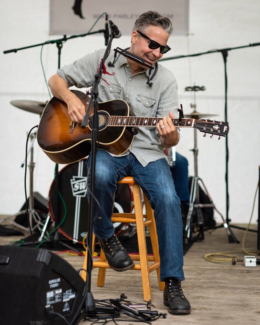 A man seated on stage on a stool playing guitar with a harmonica in a rack around his neck. He is wearing sunglasses, smiling, and looking down.