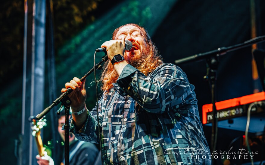 A man with rusty-colored beard and long hair closes his eyes and sings soulfully into a microphone