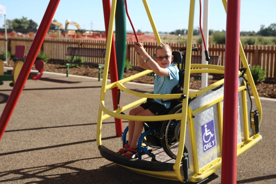 A young boy in a blue polo shirt and black shorts sitting in a wheelchair holds up his hands and pulls on a red rope. He is sitting in a yellow gondola-style bucket that has a blue "wheelchairs only" sign on it and acts as a swing. The wheelchair rolls into the structure and then the ramp folds up to be a wall of the swing. 