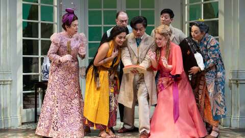 A cast of actors during a play all looking together at a page in a book with different expressions