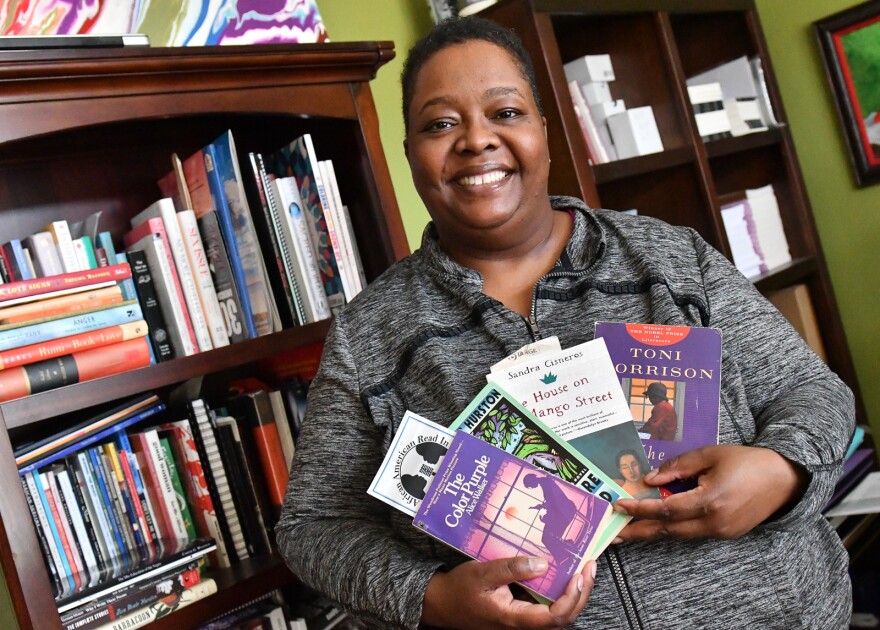 A woman stands in front of bookcase holding copies of "The Color Purple," "Their Eyes Were Watching God," "The House on Mango Street" and "The Bluest Eye" are some of the books Natasha Ria El-Scari recommends.