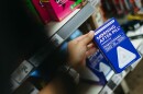 A hand holds a box of blue emergency contraceptives with the background of a convenience store shelf.