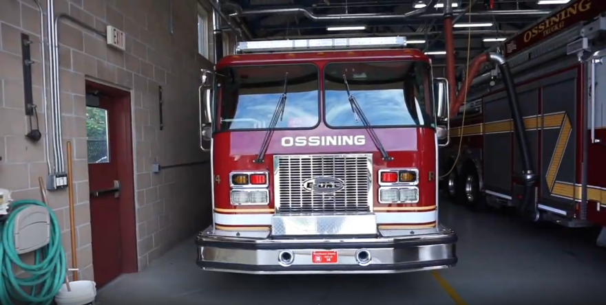 This is the front of a fire truck with the name of the town, Ossining, across the front. The fire truck is in a brick garage with another fire truck next to it. 