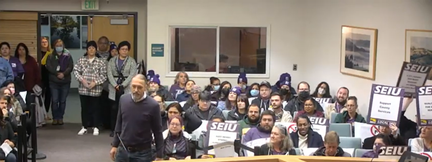 A man approaches a microphone in a crowded meeting room, full of people wearing purple shirts and holding signs that read "SEIU," representing a labor union.