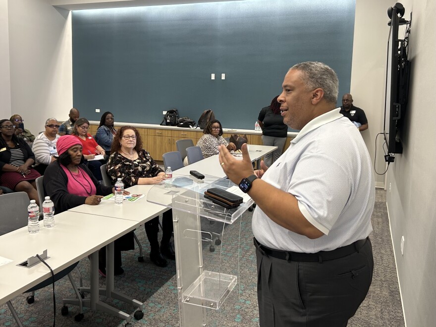 A photo of Tarrant County Commissioner Roderick Miles Jr., a Black man with short salt and pepper hair and wearing a white polo and black pants, speaks at a podium in front of a small meeting room filled with people listening to his remarks.