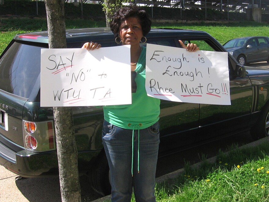 Sheilla H. Gill had been a counselor at a D.C. high school for 32 years before being fired last year. She held signs of protest near a press conference on Wednesday where Rhee and the teachers union declared they had reached a tentative agreement.