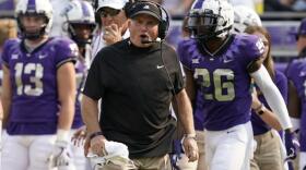  TCU head coach Gary Patterson wears a black  t-shirt on the sidelines of a game. He's surrounded by TCU players in purple jerseys and white pants.