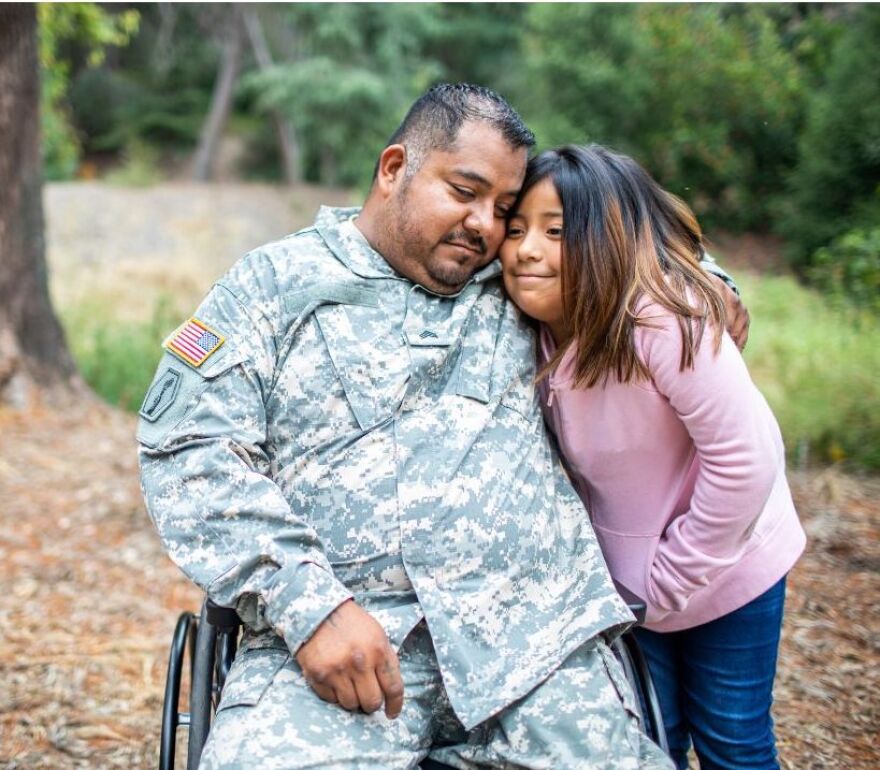 Veteran wearing camo uniform sitting in wheelchair hugs his young daughter, both with heartfelt expressions