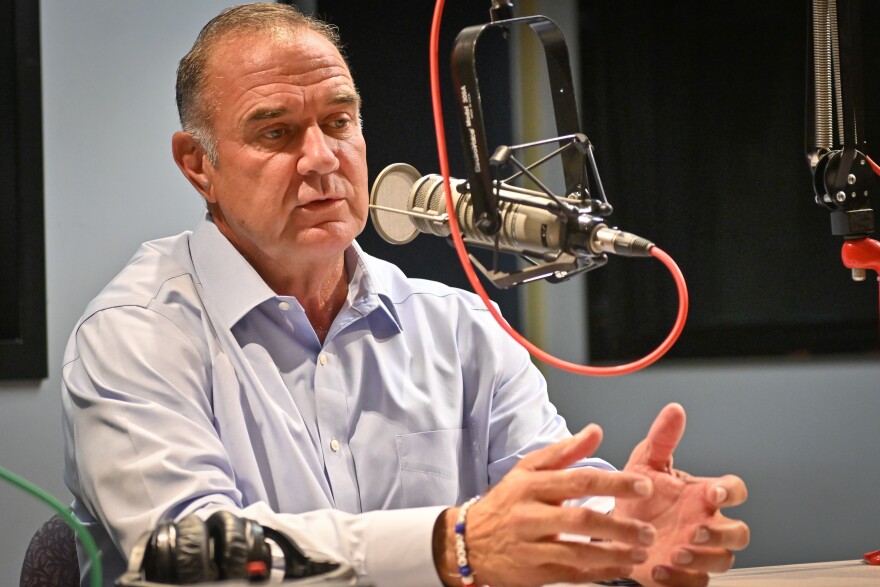 A man inside a radio station sits behind a microphone. He is talking and gesturing with both hands.