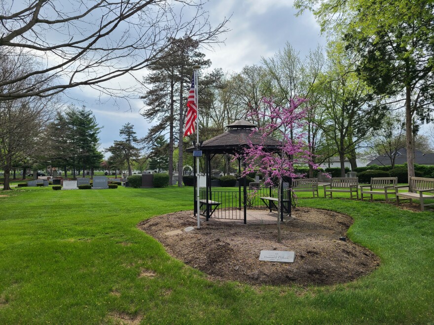 a gazebo in a cemetery with a small redbud tree blooming and a USA flag. an arc of benchs flank one side of the gazebo