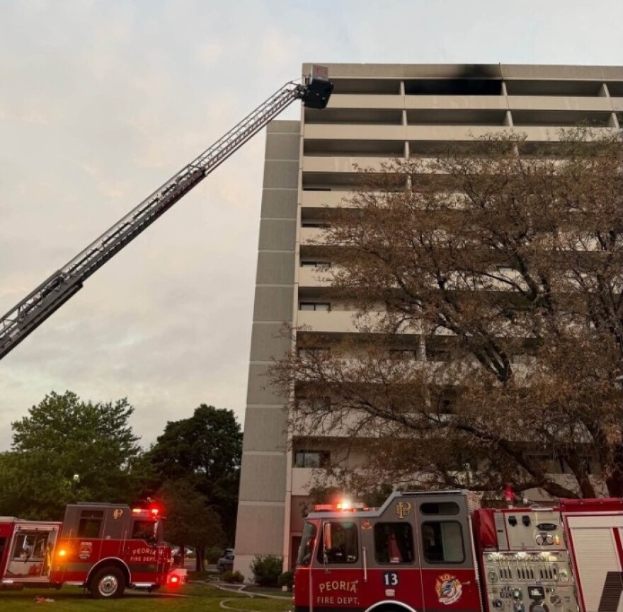 Fire trucks with extended ladder respond to a fire on the upper floor of a multi-story building, with black smoke visible from the windows. Trees and emergency lights are also visible in the scene.