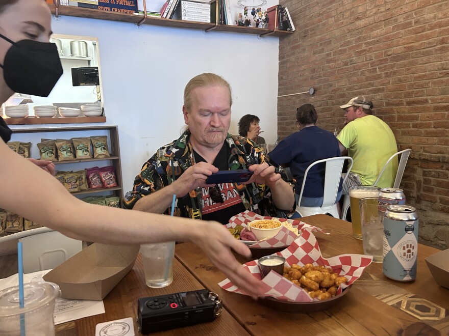 A man takes a photo of his food with his phone at a restaurant table while a masked server reaches across to place a dish. Other diners sit in the background against a brick wall.