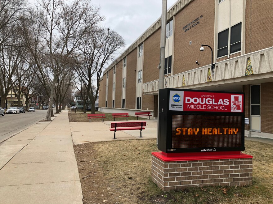 On the sidewalk outside a school building, an electric school sign says, "STAY HEALTHY" under the label "Andrew Douglas Middle School." 