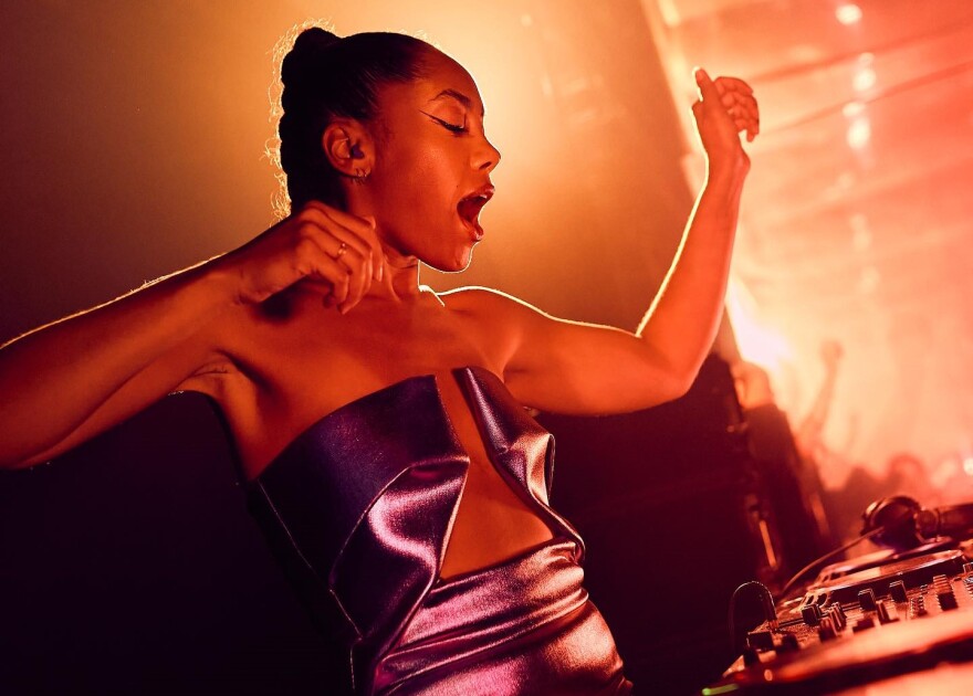 A woman in a shiny, low-cut top sings and dances while operating a sound board in a club setting.