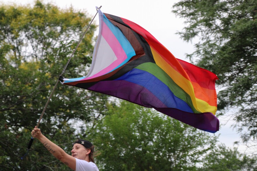  A person waves a progress pride flag in the air. The flag has a triangle made of stripes on the left side that has the colors white, pink, blue, brown and black. Horizontal stripes are red, orange, yellow, green, blue and purple.