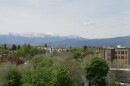 A wide view of Missoula, Montana, shows leafy green neighborhoods and low-rise buildings beneath snow-capped mountains under a hazy spring sky.