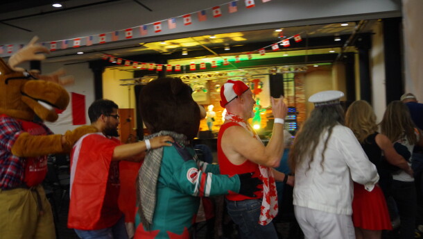 A line of people are pictured in a room where the ceiling is decorated with dozens of tiny american and canadian flags. The people have their hands on eachother's shoulders and hips in a conga line. In the line is a moose mascot wearing a plaid flannel, a man draped in a canadian flag, a beaver mascot wearing a hockey jersey, a man in a canadian flag hat with red moose ears, and a woman in a white jean jacket and white jean pants wearing a white conductor's hat. 