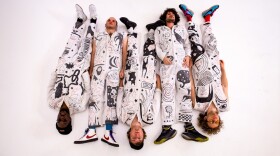 Five men wearing jumpsuits with drawings on them lay down on a white floor and look at the ceiling.