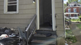 A rear entrance to a single family home. The door and a window are wide open and trash is piled on the ground.