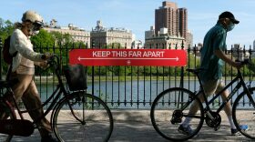 People wearing protective masks walk their bicycles past a social distancing sign reading "KEEP THIS FAR APART" at Jacqueline Kennedy Onassis Reservoir in Central Park.