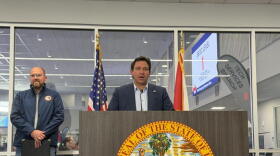Man with brown hair and a dark suit jacket standing behind a podium with a yellow state seal, in front of a glass window and a man in a blue windbreaker to the left