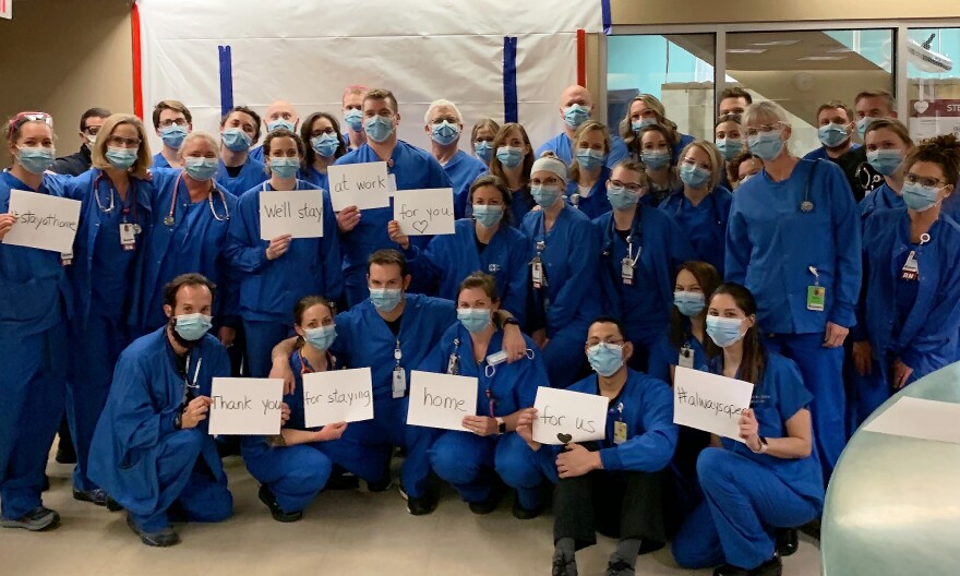 Health care providers at St. Mary's stand together with paper signs that say, "stay at home, we'll stay at work for you, thank you for staying home for us, always open"