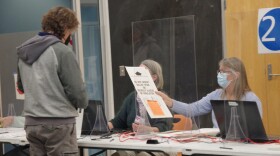 A man in hooded sweatshirt and cargo pants receives a ballot from a poll worker wearing a surgical mask.