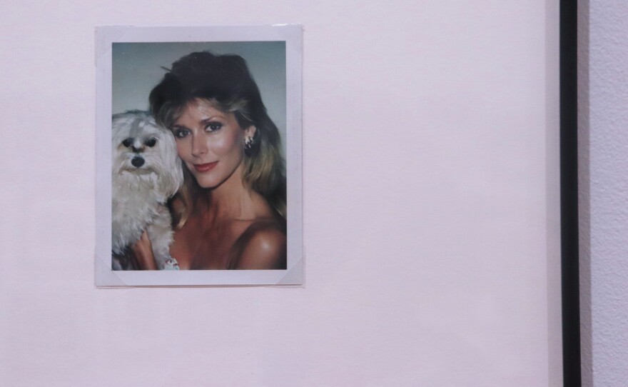 A small square photo sits against a white wall. The photo shows a blonde woman holding a white dog. She smiles softly at the camera. 
