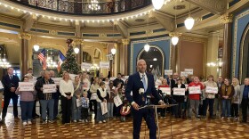 a man speaks at a podium in front of a few dozen people and a Christmas tree