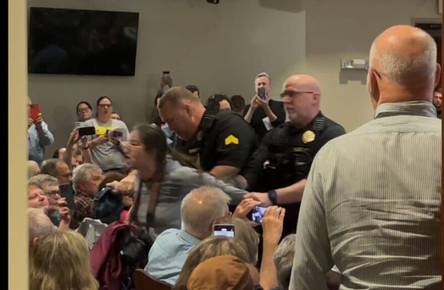 A women in a grey sweatshirt is restrained by two police officers leading her out of a room of people.
