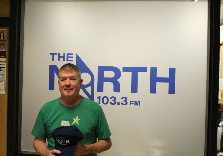 A man holding a baseball cap that says the North 103.3 in front of a frosted window that says the North 103.3 FM