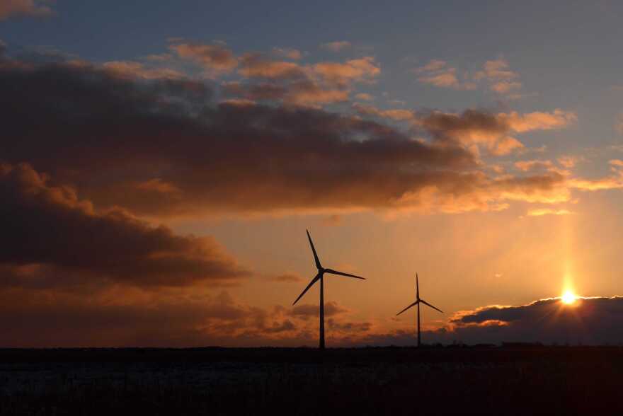 Two wind turbines are set in shadow as a sun sets in the background. 