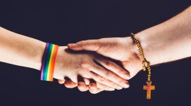 A photo of two hands shaking, one with a lgbtq+ pride band and the other with a bracelet with a cross.