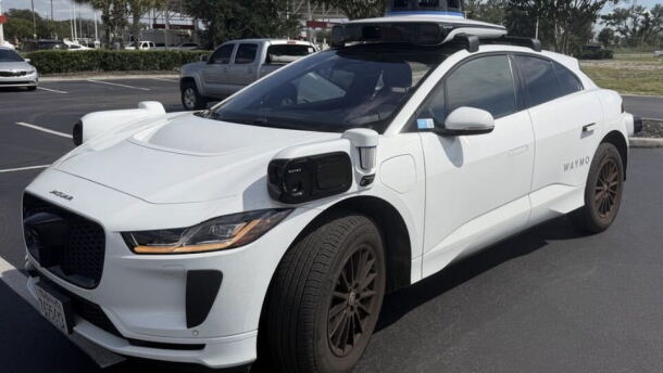 A white Jaguar car with black sensors on the front sides and on the roof sits in a parking lot in-between spots.