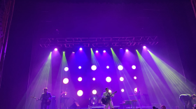  A crowd looks at a stage with a band. The frontrunner is a woman with a white dress and an acoustic guitar. Purple lights shine on the musicians.