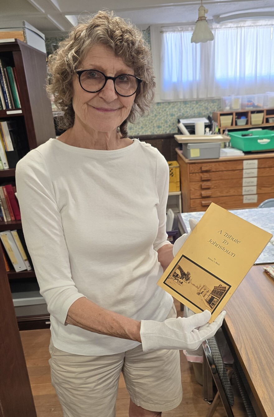 A woman wearing a cream-colored shirt, khaki shorts and black-rimmed glasses holds up a small, tan-colored book. The front cover of the book bears the words "A Tribute to Johnstown by Rebecca S. Healy" in black text. A black-and-white photo on the cover features Johnstown's early downtown area. Horse-drawn carriages travel on a dirt road lined with shops.
 