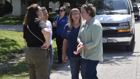 Four women standing next to each other in a semi circle on a residential street and in front of a white van.