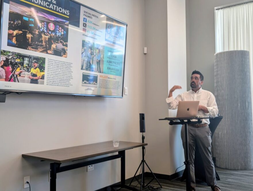 A man in a white button-down shirt and grey pants gives a presentation. There's a laptop on a podium in front of him and a display screen to the left.