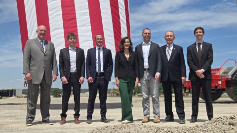 A group photo of seven people, including Michigan Governor Gretchen Whitmer, standing in a line at an outdoor construction site. They are dressed in business professional and business casual attire. In the background, a massive American flag hangs vertically behind them against a partly cloudy blue sky, alongside heavy construction machinery. In the foreground is a large pile of gravel.