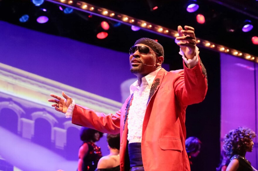 Black man wearing sunglasses and peach suit jacket singing on stage with his arms out