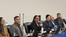 Seven councilmembers sit at a table in front of computers and microphones 