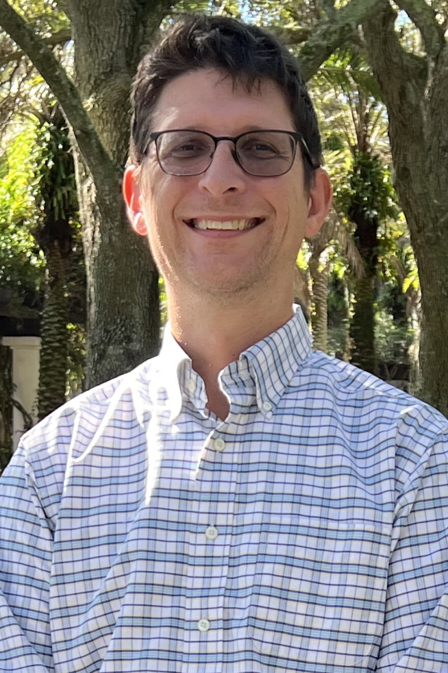 Man with a white and blue patterned shirt, brown hair and glasses smiling into the camera