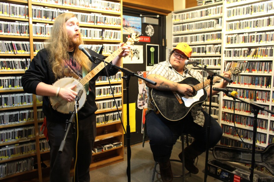 A standing person on the left plays banjo and sings into a microphone while a seated person on the right plays guitar. They are in a radio studio whose walls are lined with shelves full of CDs and an emergency exit door covered in band posters.