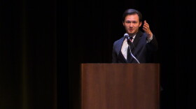 A young man in a suit extends his arm out to the crowd under a spotlight with a black background 