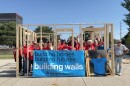 Volunteers in red T-shirts stand inside the frame of a square house, holding a banner that reads "building homes, building futures, building walls."