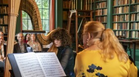 In the foreground, flutist Kathy Karr wearing a yellow top with her back to camera, across from Grace Roepke playing harp in a library
