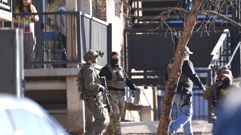 Multiple people in camouflage and tactical gear carry rifles outside of a large building.